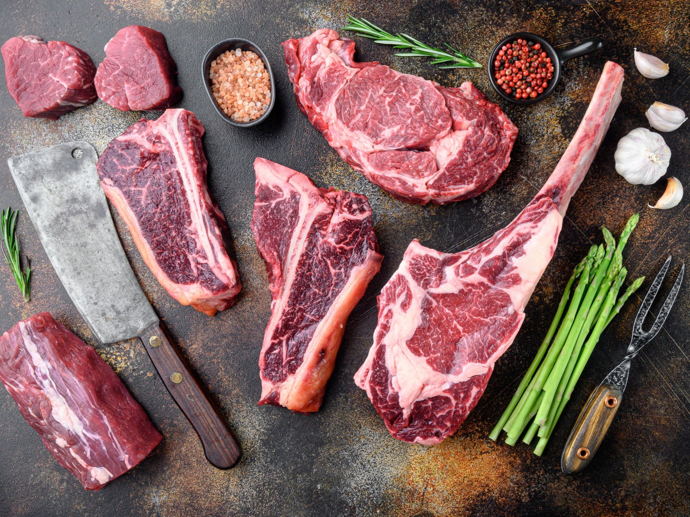 High-quality cuts of beef including ribeye, tenderloin, and tomahawk steak arranged on a rustic surface, accompanied by a cleaver, fresh rosemary, pink salt, mixed peppercorns, garlic cloves, and asparagus, showcasing the ingredients for a gourmet meal.