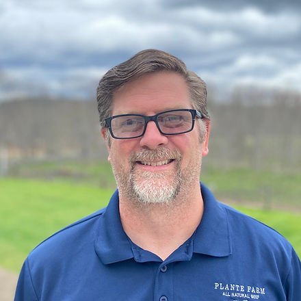 IMG_03852_edited Smiling man with glasses wearing a blue polo shirt stands outdoors in a farm setting, with green fields and cloudy skies in the background.