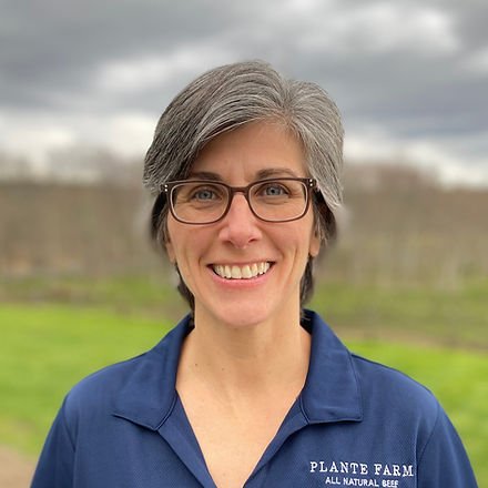 IMG_0400_edited (1) Smiling woman with short gray hair wearing glasses and a blue shirt with "Plante Farm" logo, standing outdoors in a green field under a cloudy sky.