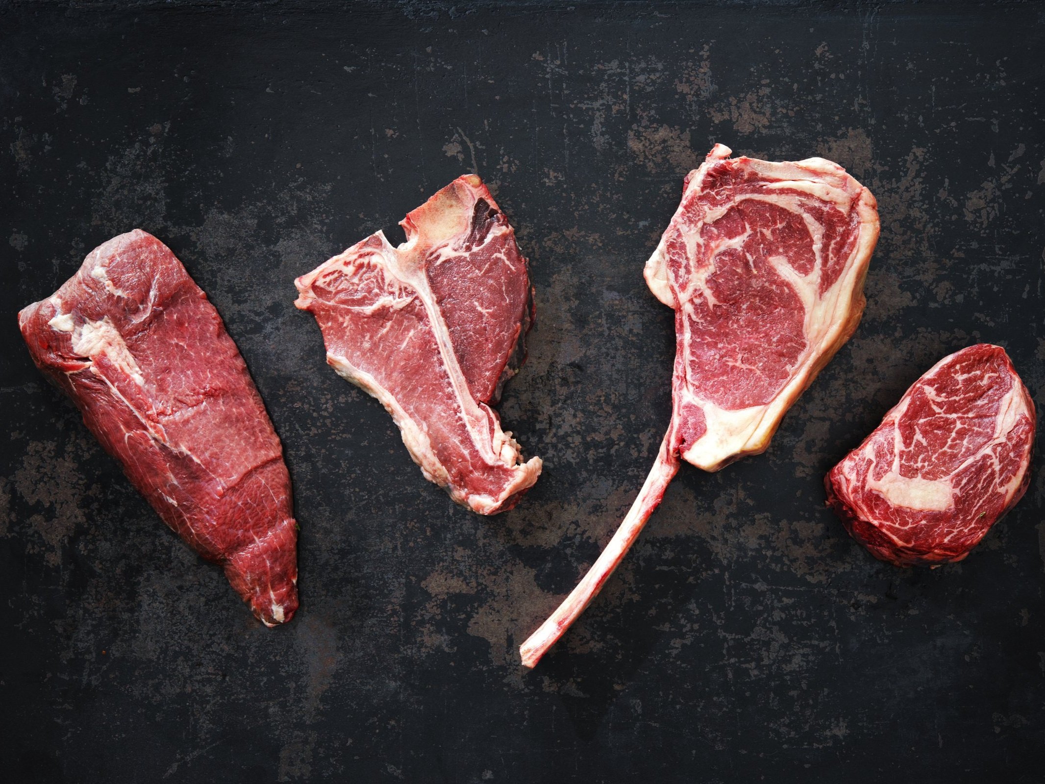 Assorted cuts of raw beef steak displayed on a dark textured surface, including ribeye, T-bone, and sirloin, showcasing marbling and freshness ideal for grilling or roasting.