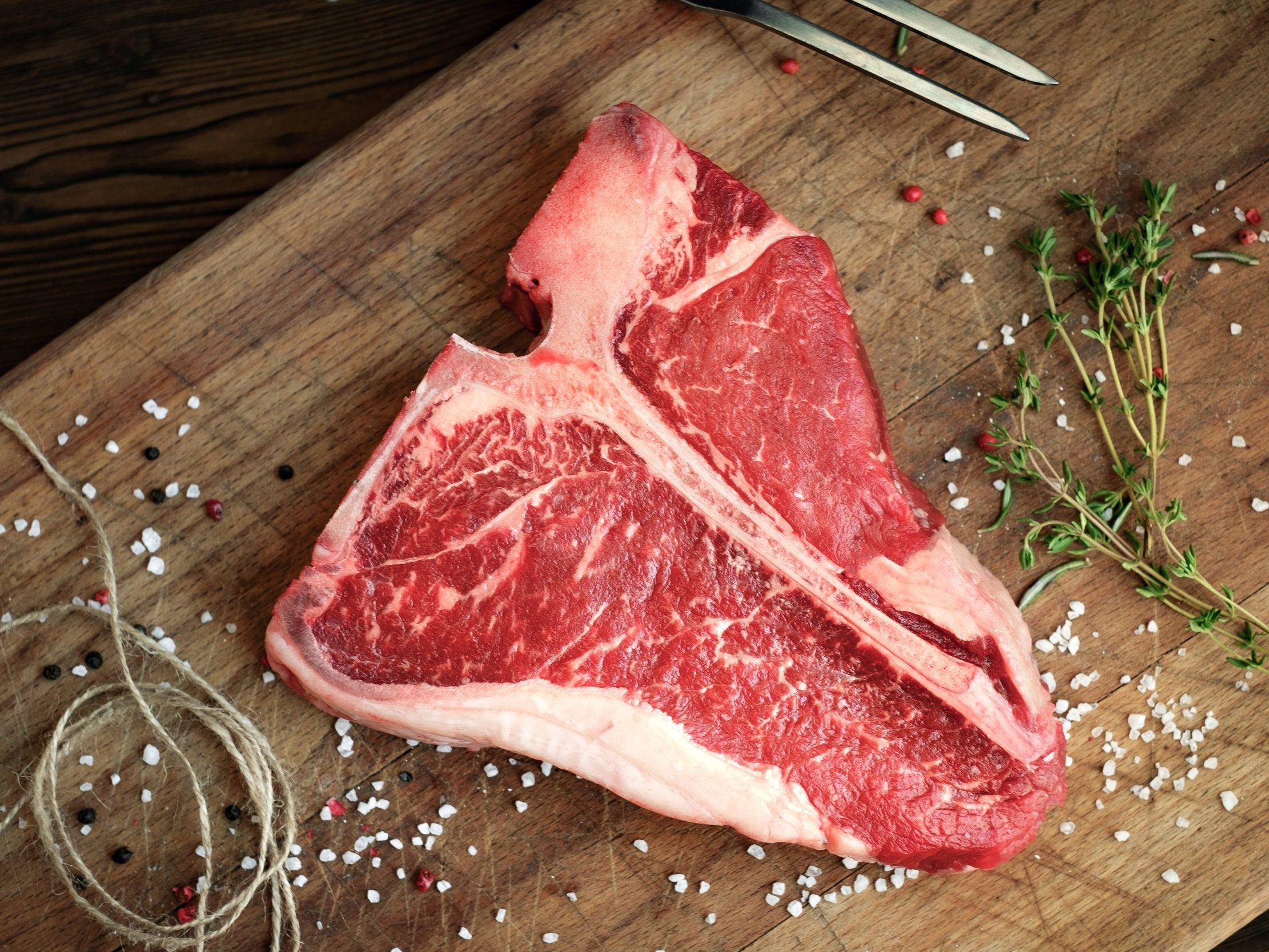 A juicy T-bone steak with marbled fat resting on a wooden cutting board, surrounded by coarse sea salt, black and pink peppercorns, and fresh thyme sprigs, ready for grilling or cooking.